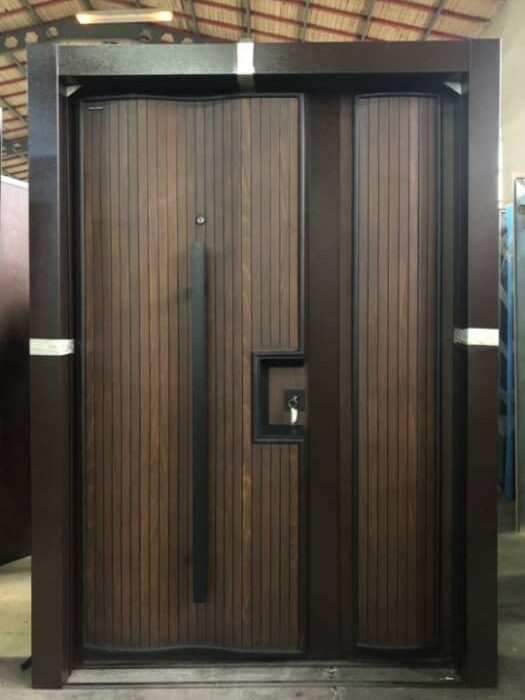 A modern double security door with a dark brown wood grain finish. The door has a black metal frame, a black handle, and a keyhole. The door is set against a concrete floor with a tiled roof visible in the background.