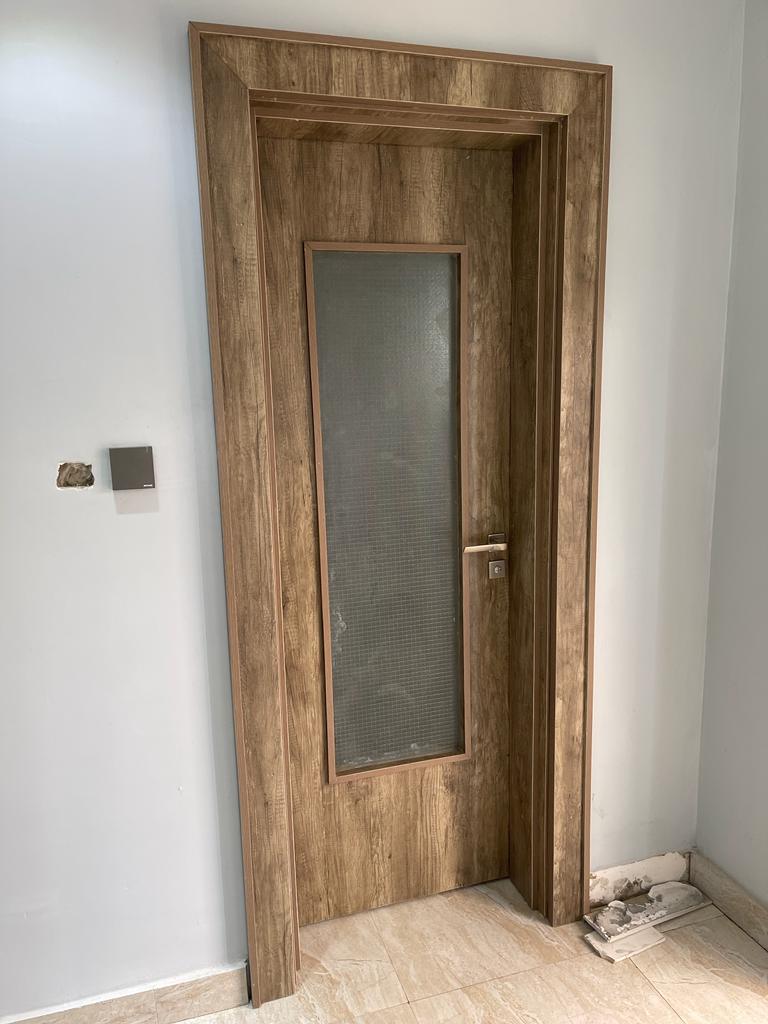 A modern Wooden interior door with a wood-look finish and a rectangular glass panel in the center. The door has a silver handle and is set against a white wall.