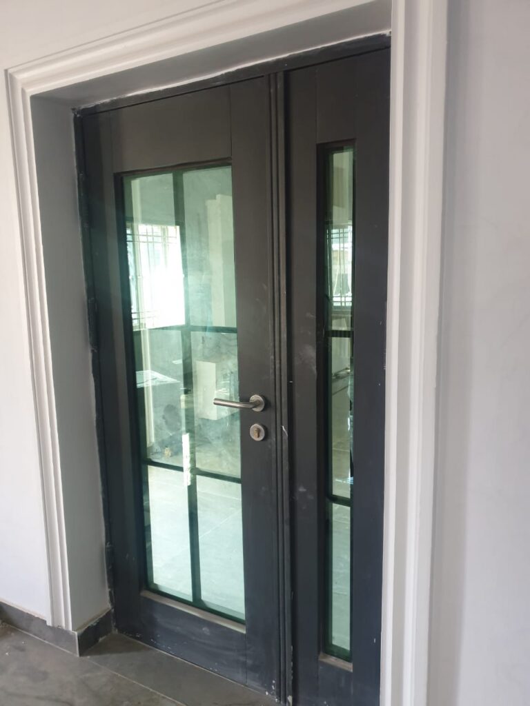 A pair of modern double doors with a black finish, featuring rectangular glass panels and a silver handle. The doors are set against a white wall with a grey floor.