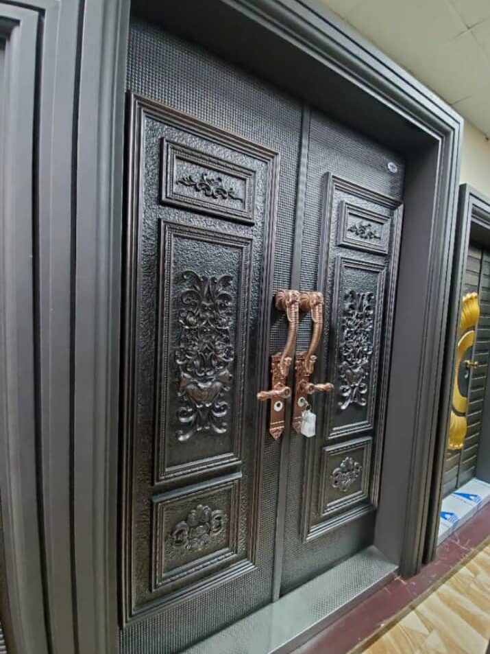 A pair of grand double security doors with a dark bronze finish, featuring intricate floral and vine patterns embossed on the metal surface. The doors have ornate handles and a peephole.